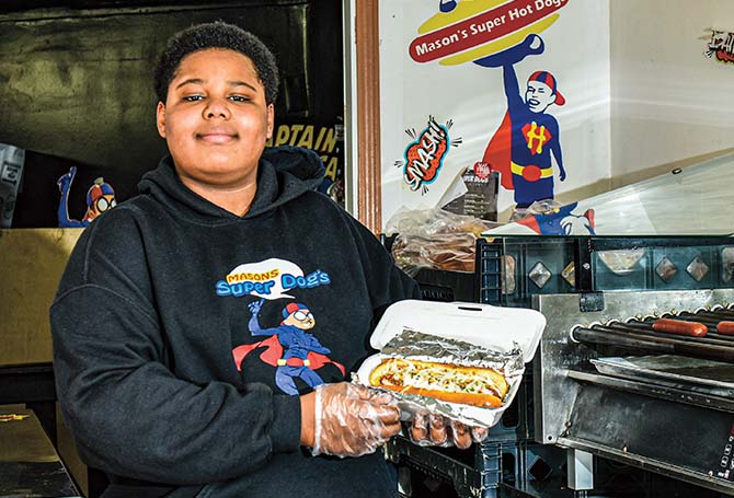 a boy wearing a sweatshirt with a Mason&apos;s Super Dogs logo and holding up a hot dog