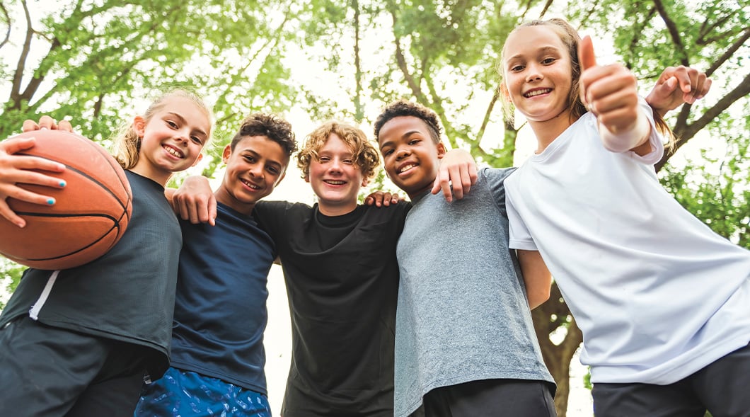 Five children stand together outdoors, one holds a basketball and another gives a thumbs up