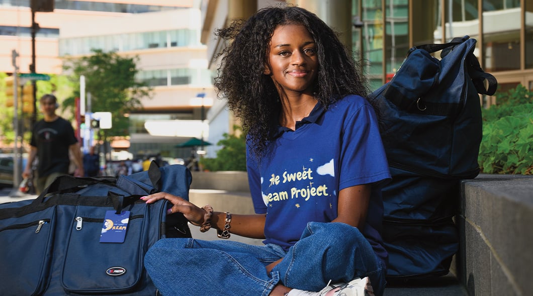 A young woman sits outdoors with duffel bags, wearing a Sweet Dream Project shirt