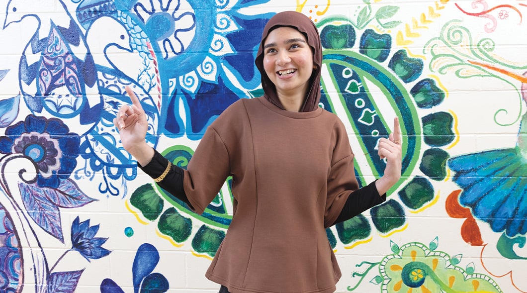 A young woman stands in front of a colorful floral mural with both index fingers raised