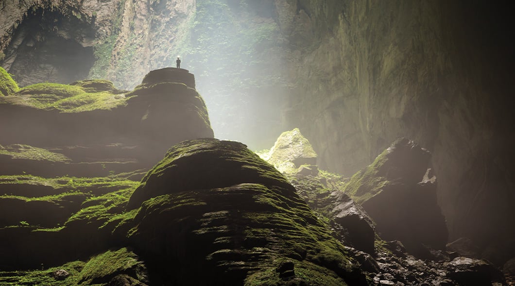A person stands atop a moss-covered rock formation inside a vast cave lit by natural light from above