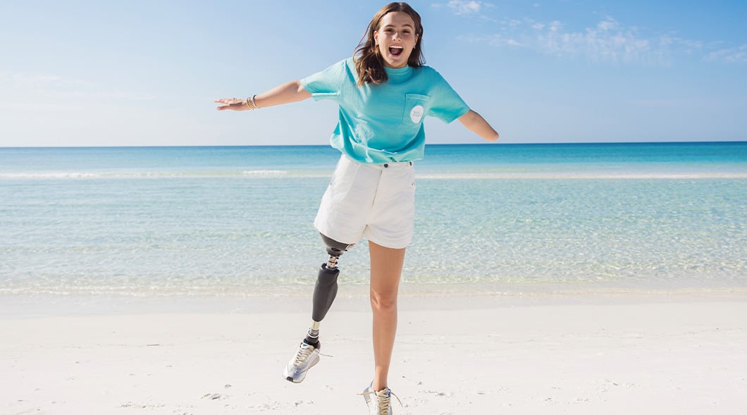 A young woman with a prosthetic leg jumps with arms wide open on a sunny beach