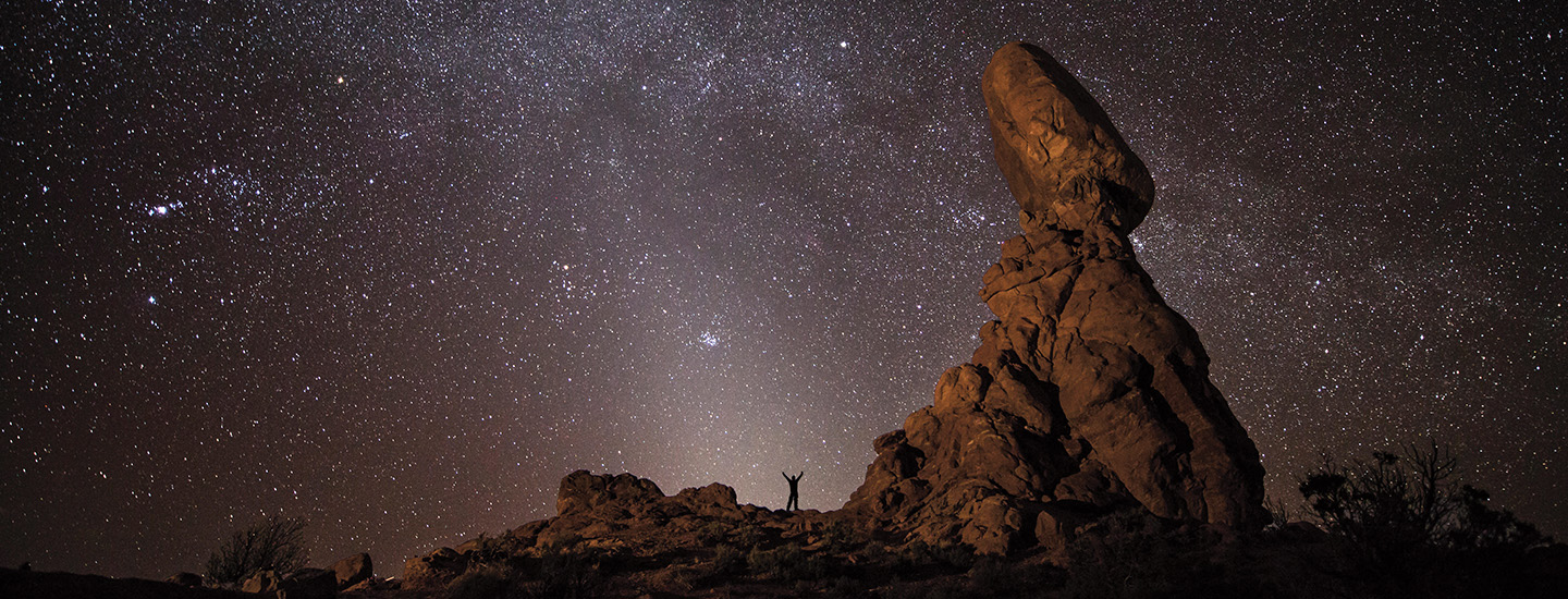 A person standing under the starry sky next to large rock formation in the middle of the desert