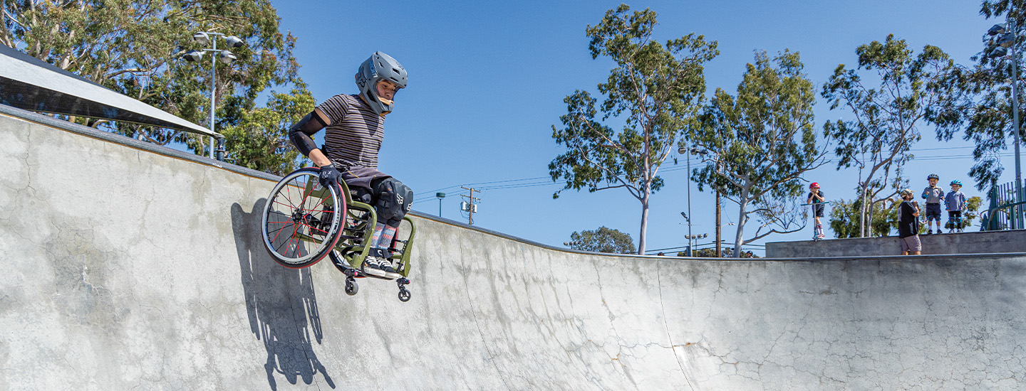 A boy in a wheelchair and helmet dropping into a skate park&apos;s pool