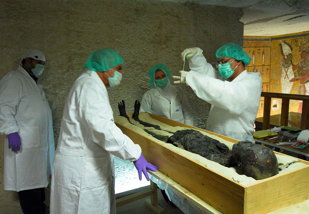Four scientists standing next to an unwrapped mummified body as they begin to examine it